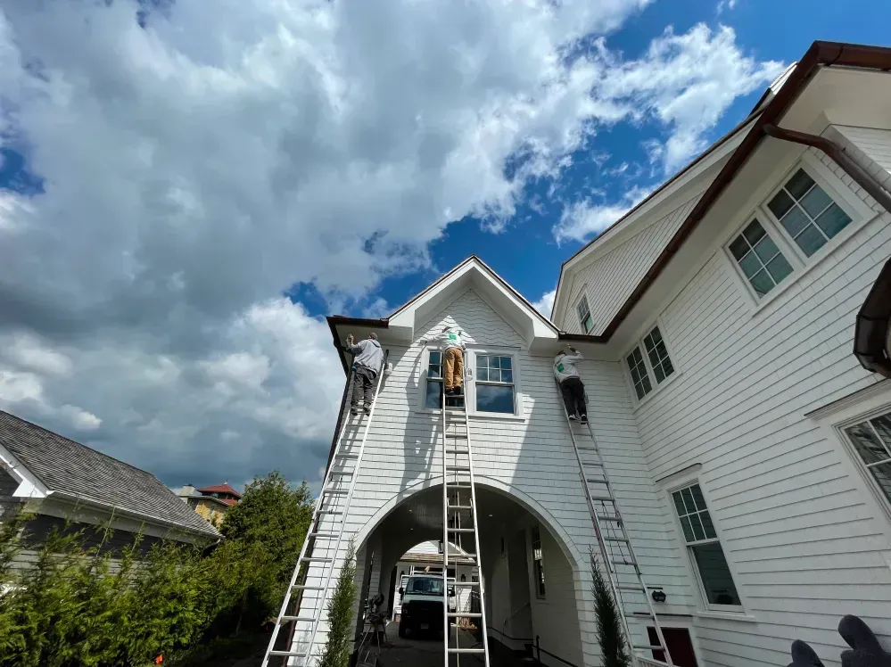 Three people on ladders painting a white house with blue sky and clouds.