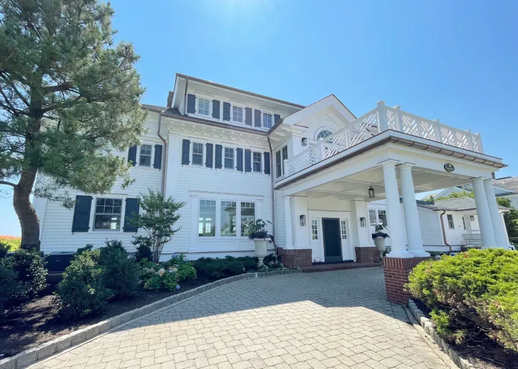 White house with columns, brick accents, and a paved driveway under a blue sky.