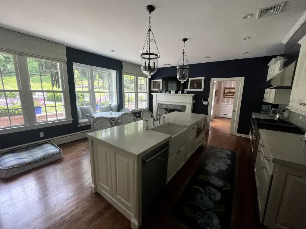 Kitchen with white island, navy walls, large windows, dining table, and two hanging lanterns.