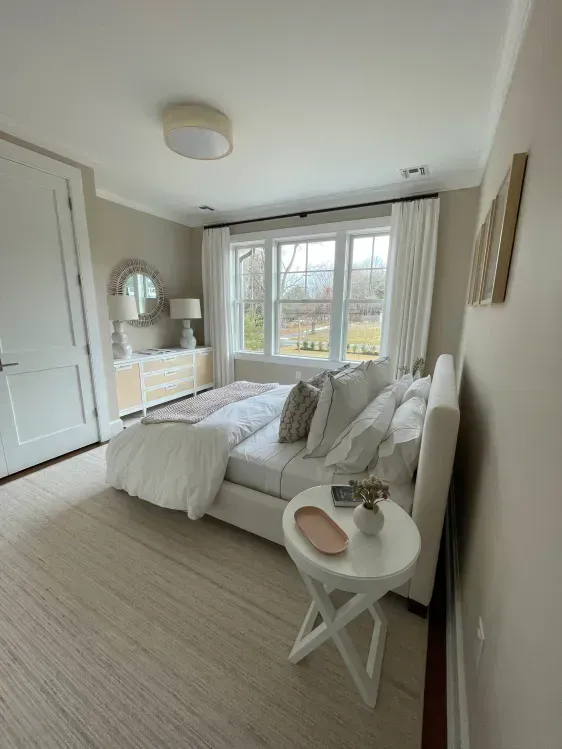 Bedroom with a white bed, beige rug, and cream-colored walls. Window with white curtains and a small white side table.