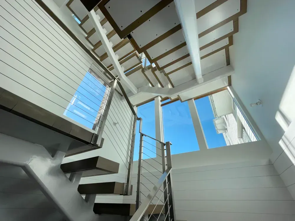 Staircase with cable railings ascending to windows and blue sky, white walls, and wood accents.