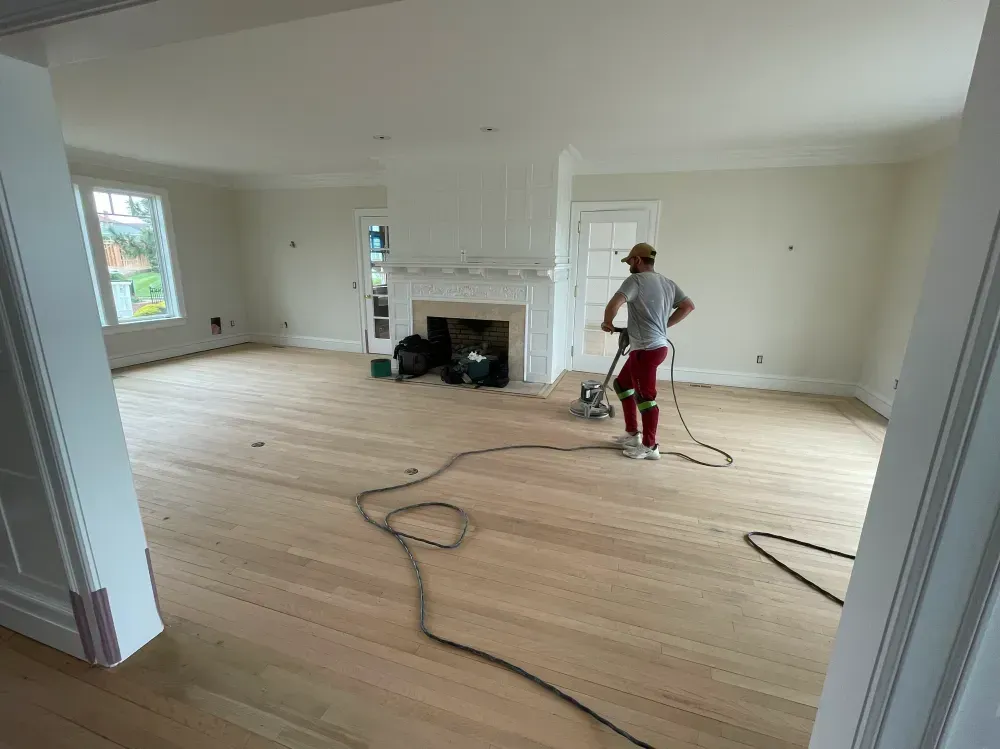 Man sanding hardwood floor in a spacious, light-filled room with a fireplace.