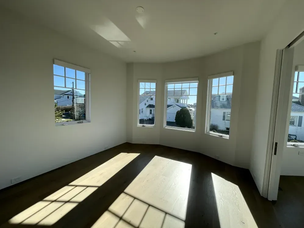 Empty room with wood floors and three-window bay looking out onto houses. Bright sunlight streams in.