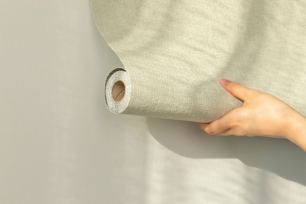 Person holding a roll of textured wallpaper against a light gray wall.