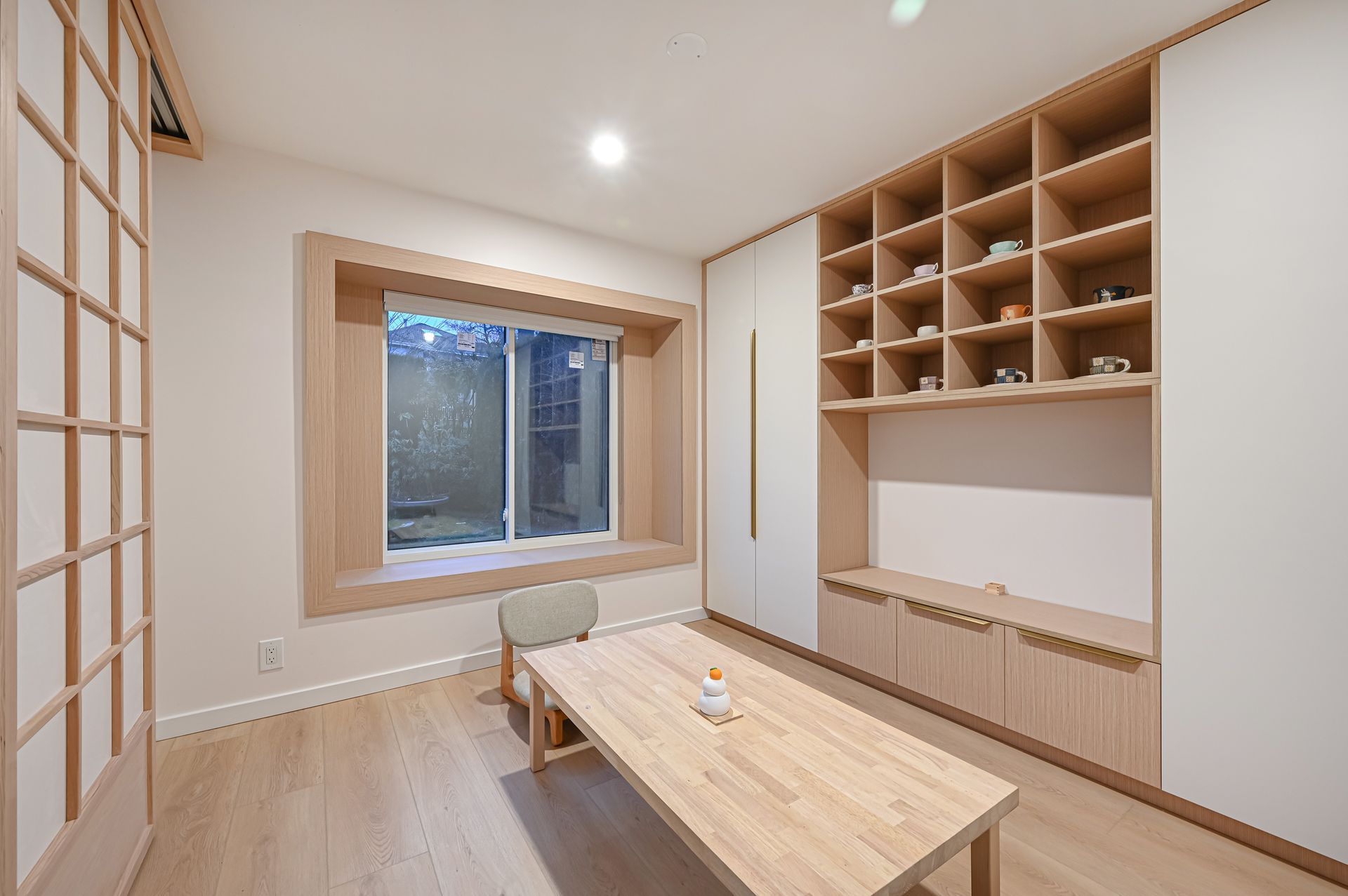 A minimalist room with light wood flooring, a central wooden table, floor chair, deep-set window, and built-in shelving.