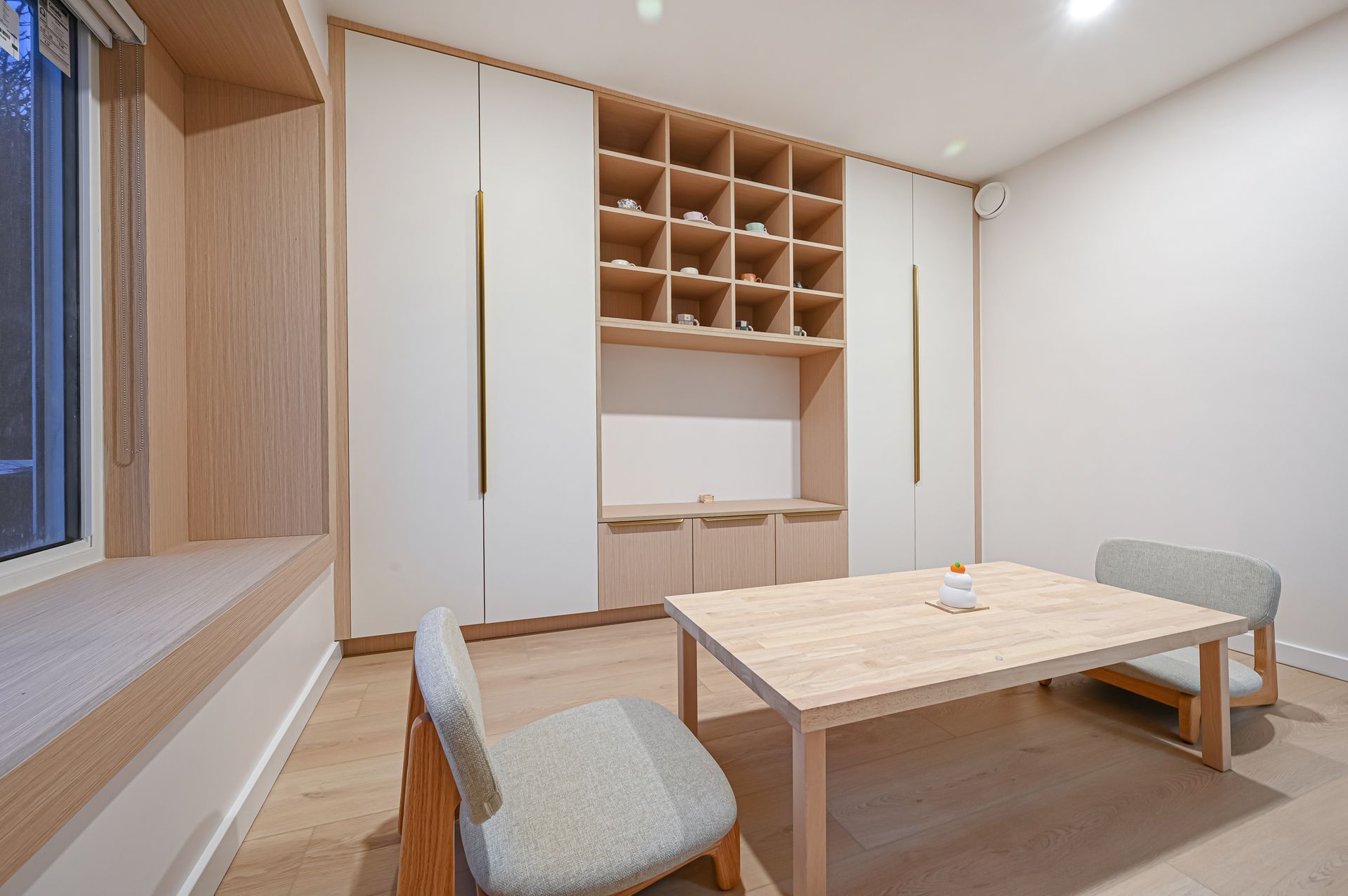 A modern, minimalist room with a light wood table, two low floor chairs, and a large custom white and wood storage unit.