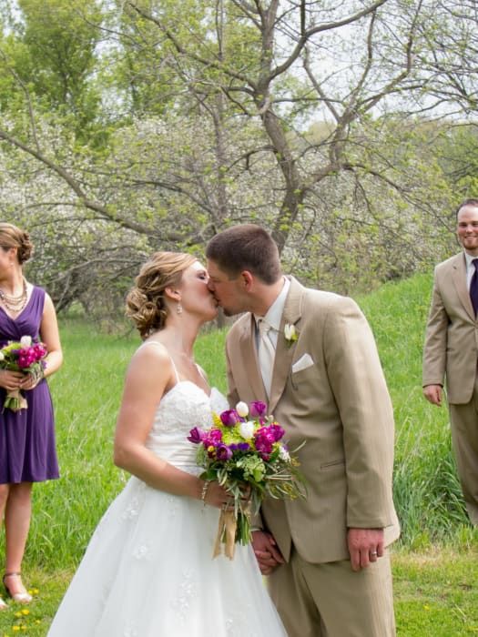A bride and groom are kissing in a field while their wedding party watches.