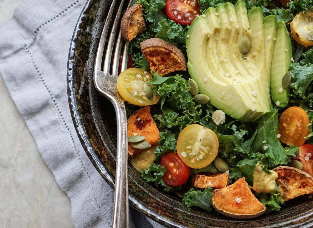 A salad with avocado , sweet potatoes , tomatoes , and kale in a bowl with a fork.