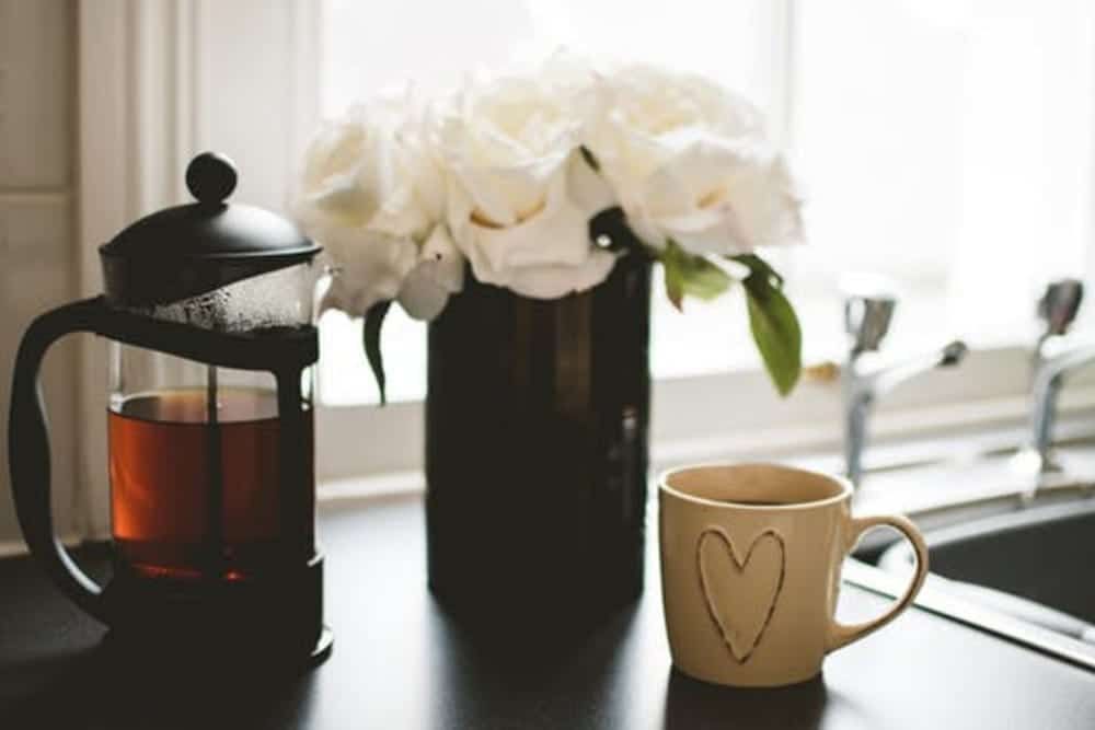 A cup of coffee , a french press , and a vase of flowers are on a kitchen counter.