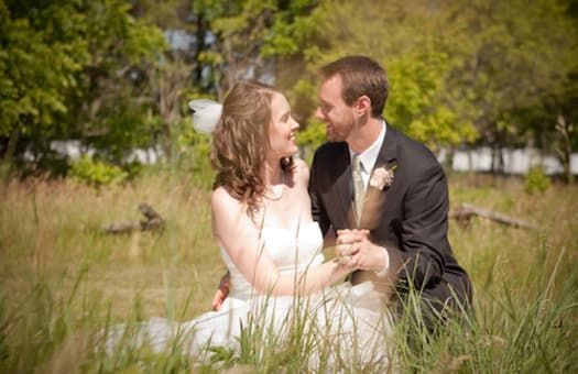 A bride and groom are sitting in the grass looking at each other.