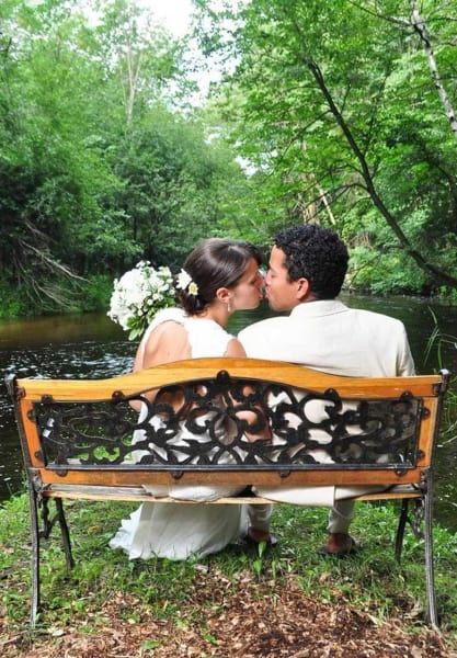 A bride and groom are kissing on a bench near a river.