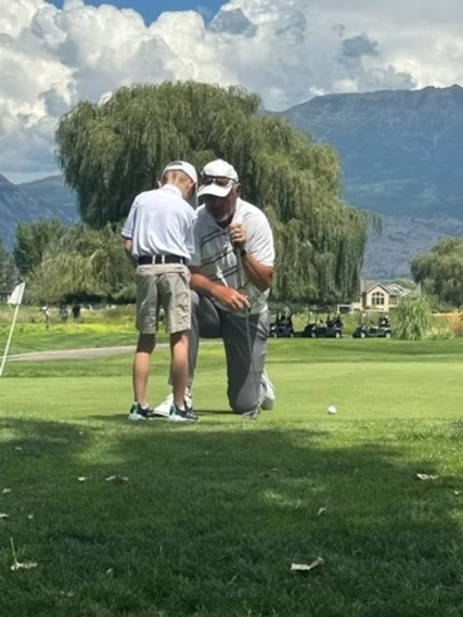 Two men are kneeling on a golf course with mountains in the background.