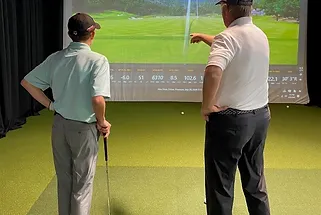 Two men are standing in front of a large screen looking at a golf course.