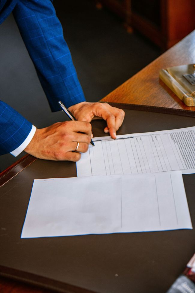 A man in a blue suit is signing a document on a desk.