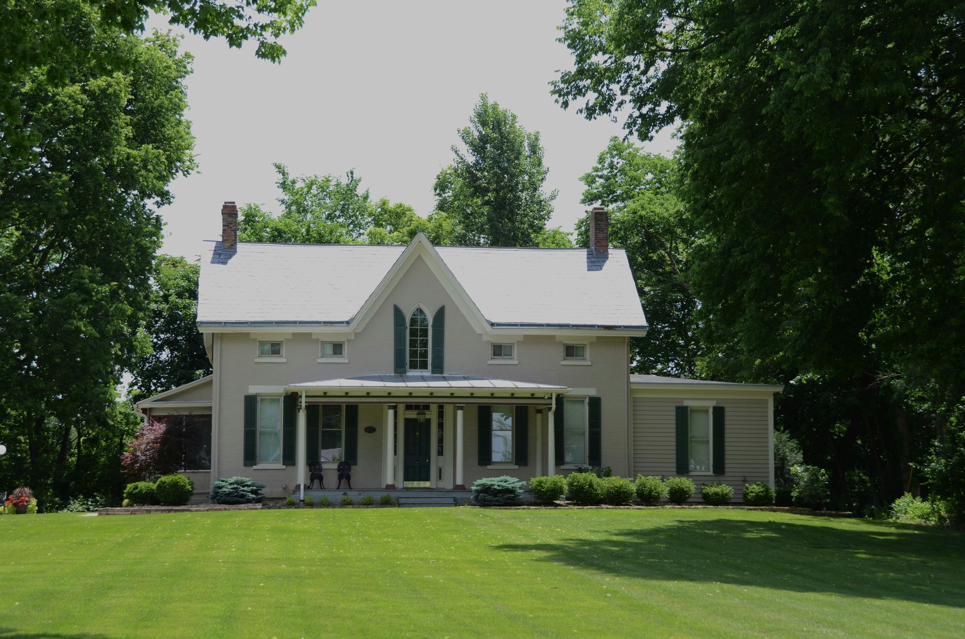 A large white house with green shutters sits in the middle of a lush green field