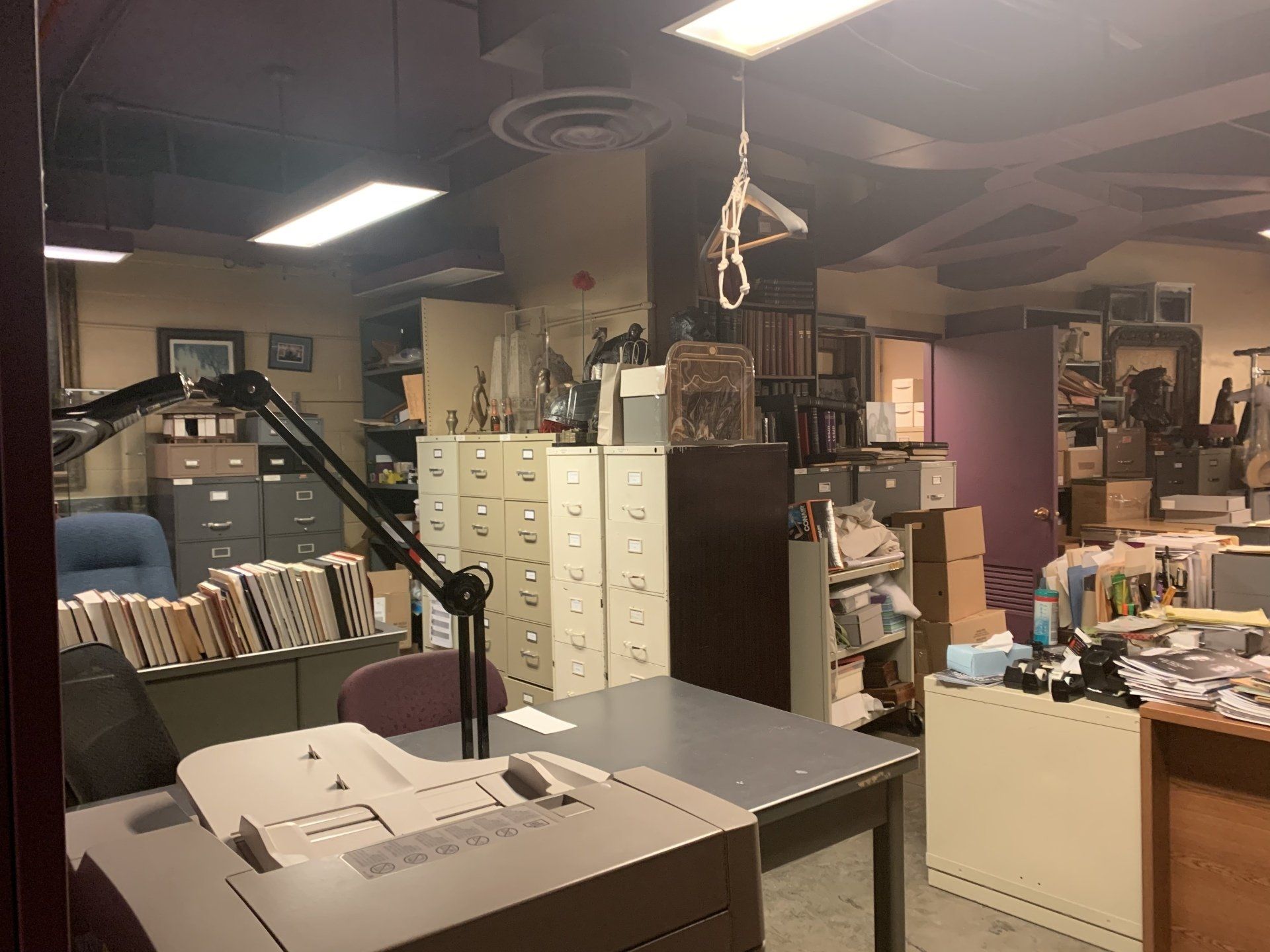 Cluttered office: gray desk, file cabinets, boxes, and a printer. Ceiling with lights and exposed pipes.