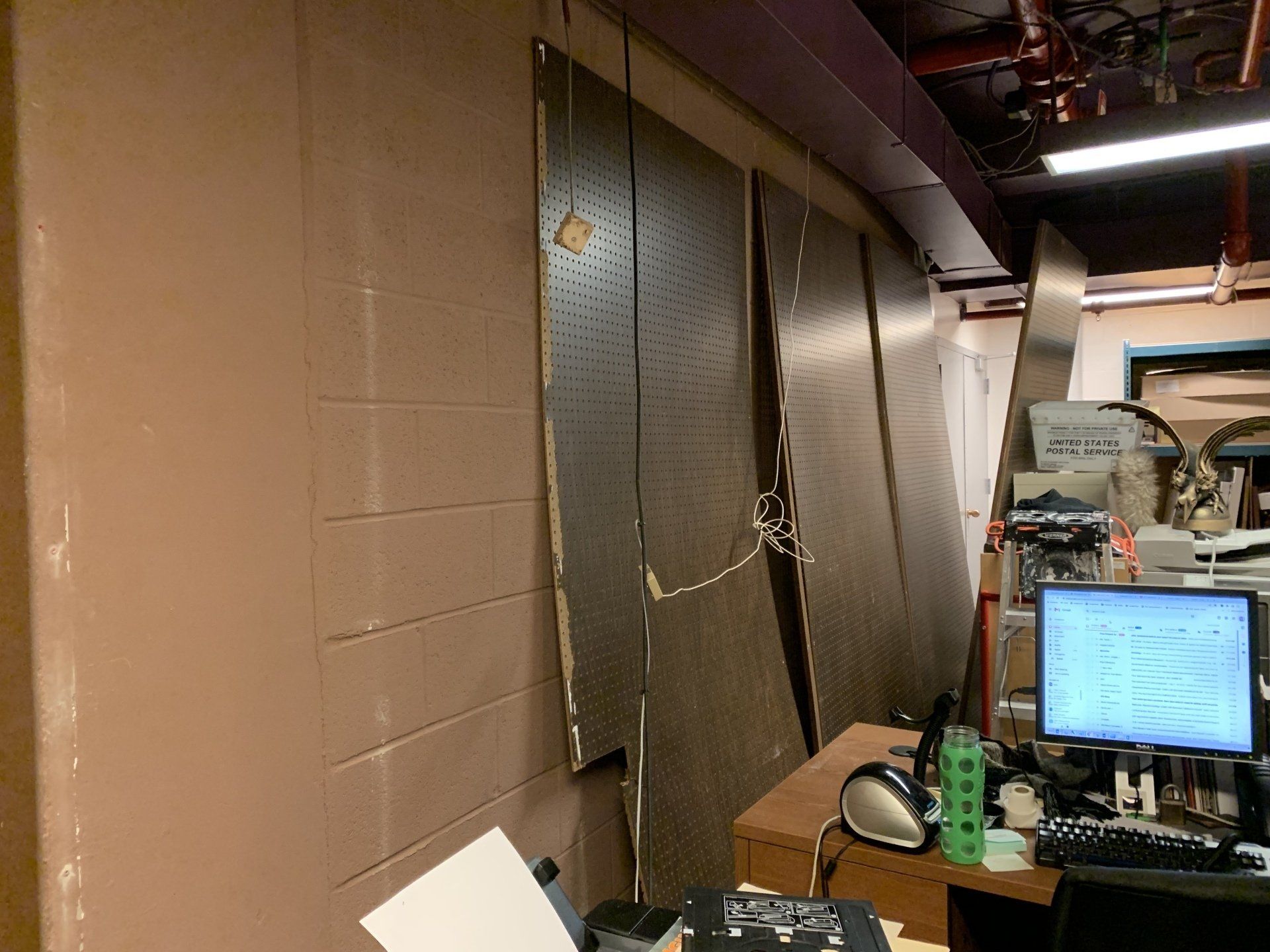 A damaged, brown wall with pegboard panels is next to a desk with a computer and equipment.