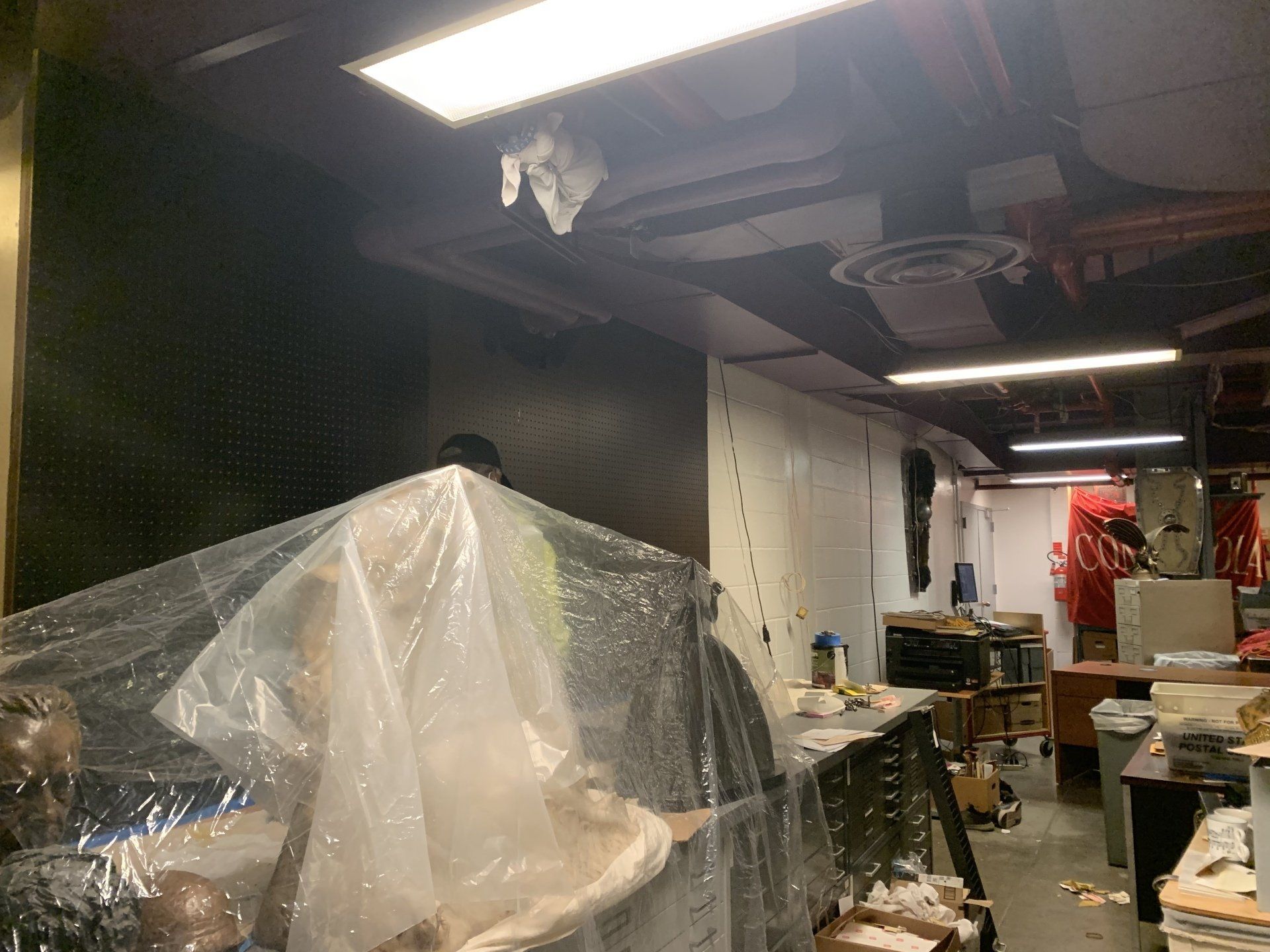 Basement workspace. Covered object in the foreground, with exposed ductwork, fluorescent lighting, and cluttered work tables.