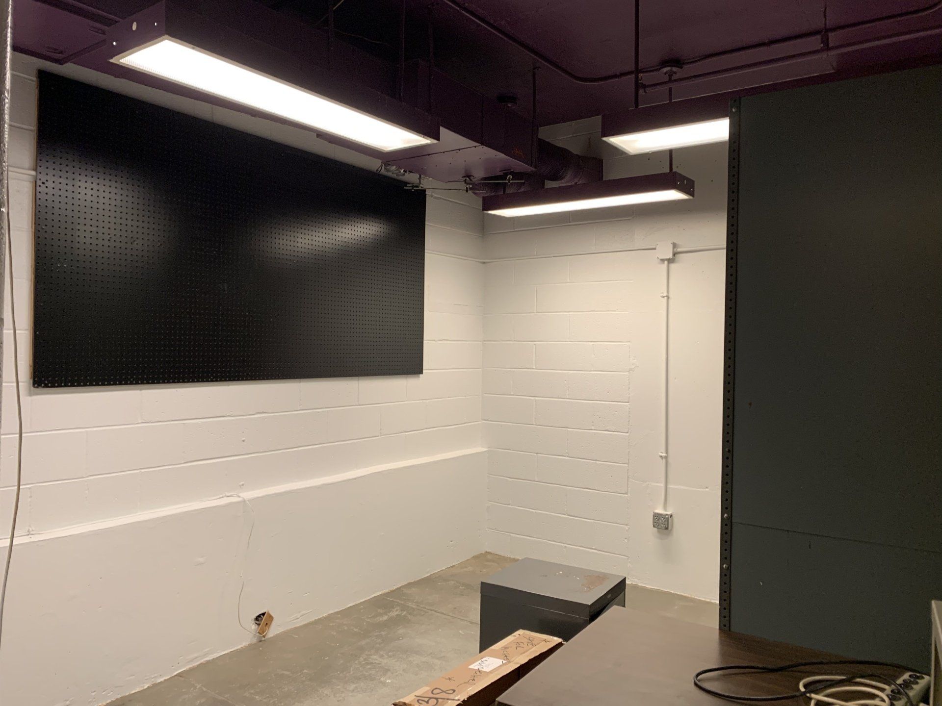 A dark-walled workshop corner with a black pegboard, concrete floor, and fluorescent lights.