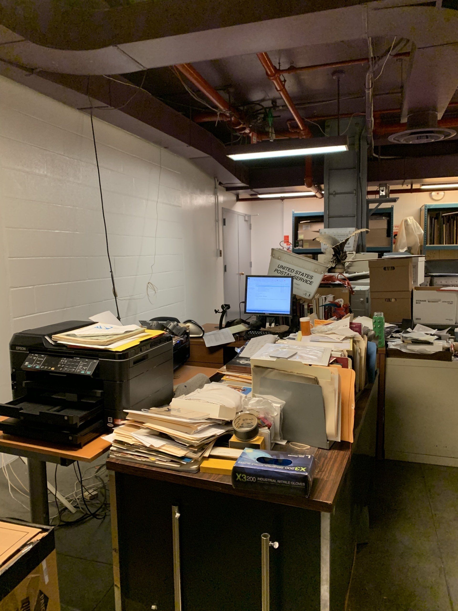 Office desk cluttered with papers, computer, and printer, in a room with exposed pipes and ductwork.