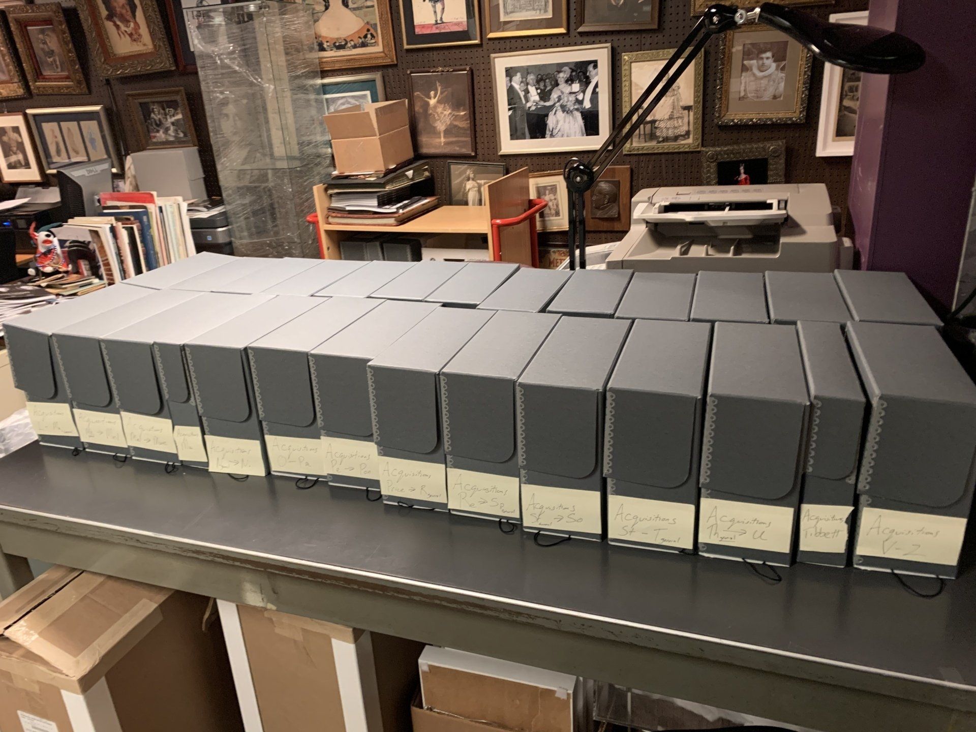 Rows of gray archival boxes on a table, against a wall filled with framed photos.