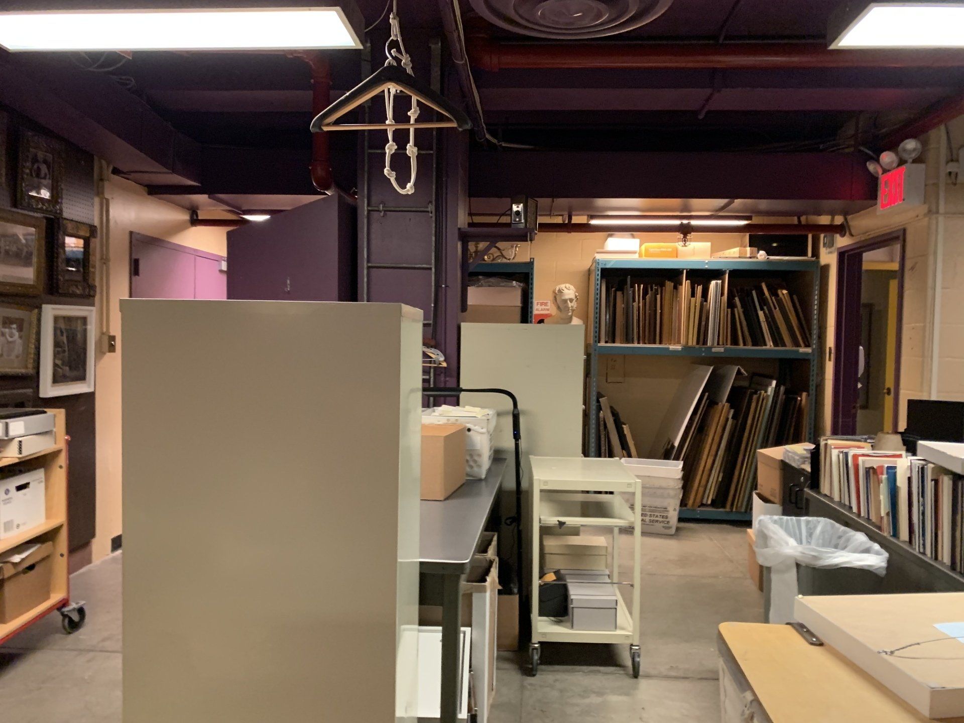 A cluttered storage room with art, shelves, and a coat hanger. Purple ceiling, gray floors.