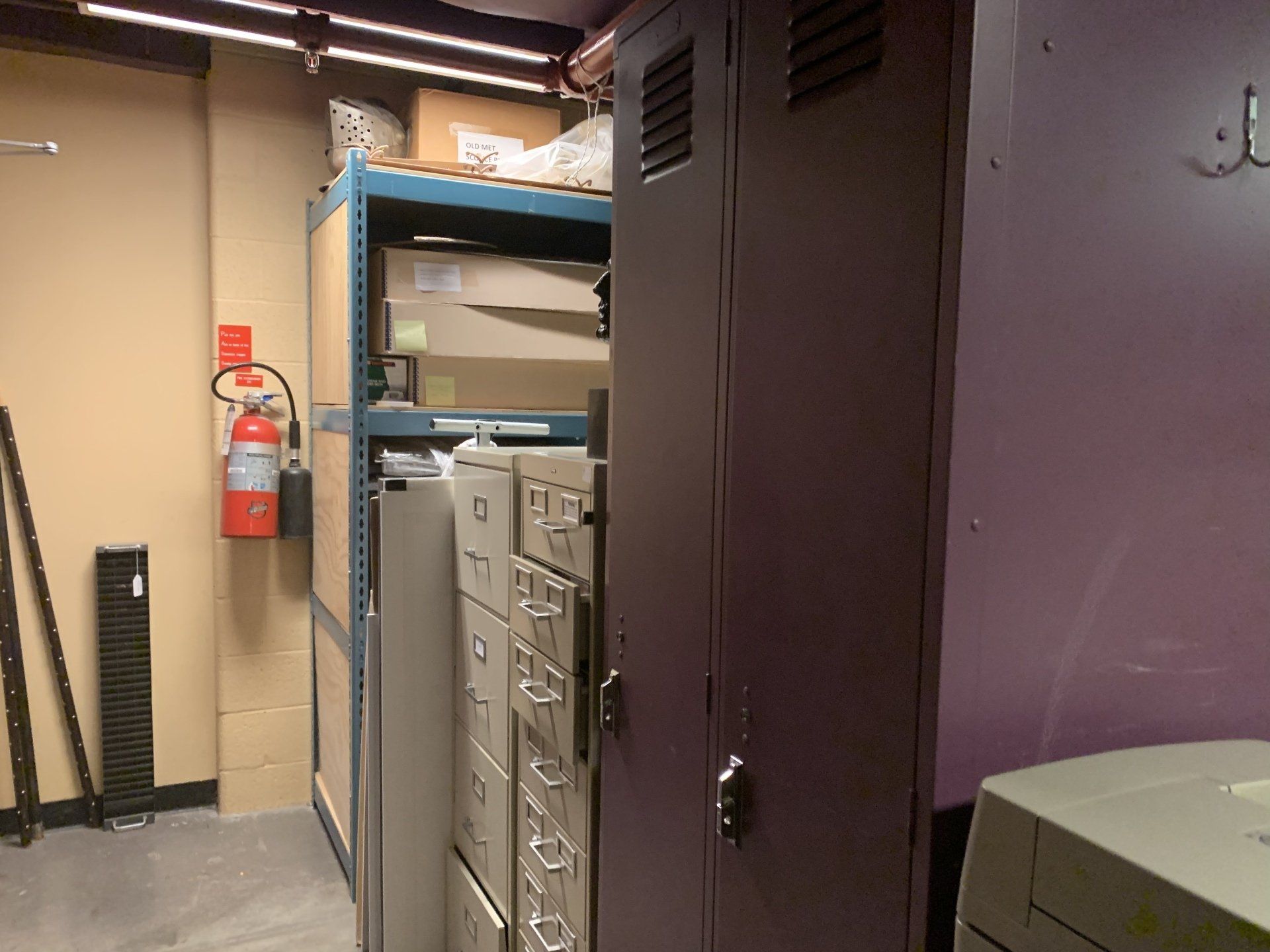 Storage room with metal shelving, filing cabinets, and a locker. A fire extinguisher is on the wall.