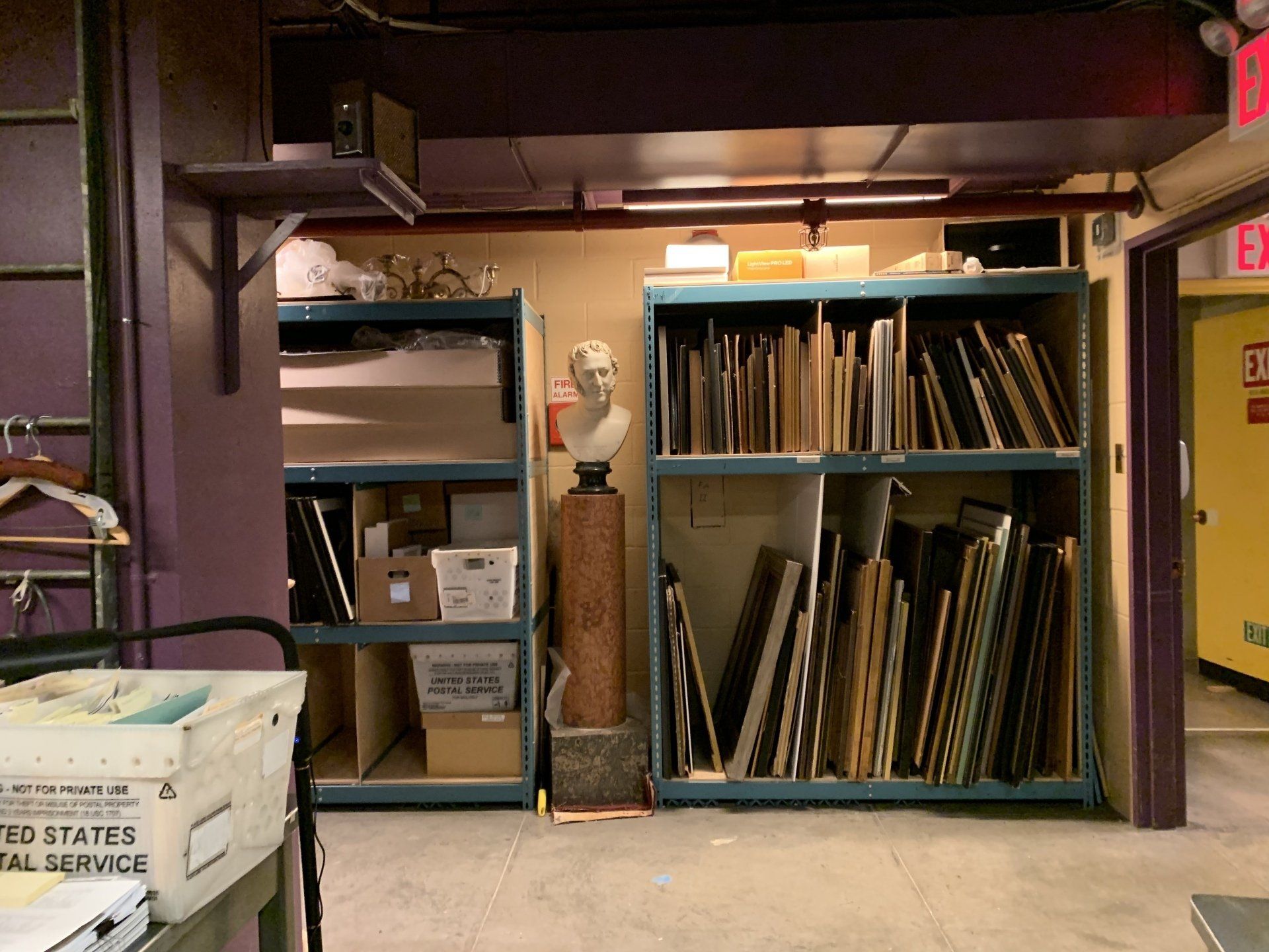 Shelves filled with artwork and supplies in a storage room; a bust statue stands on a pedestal.