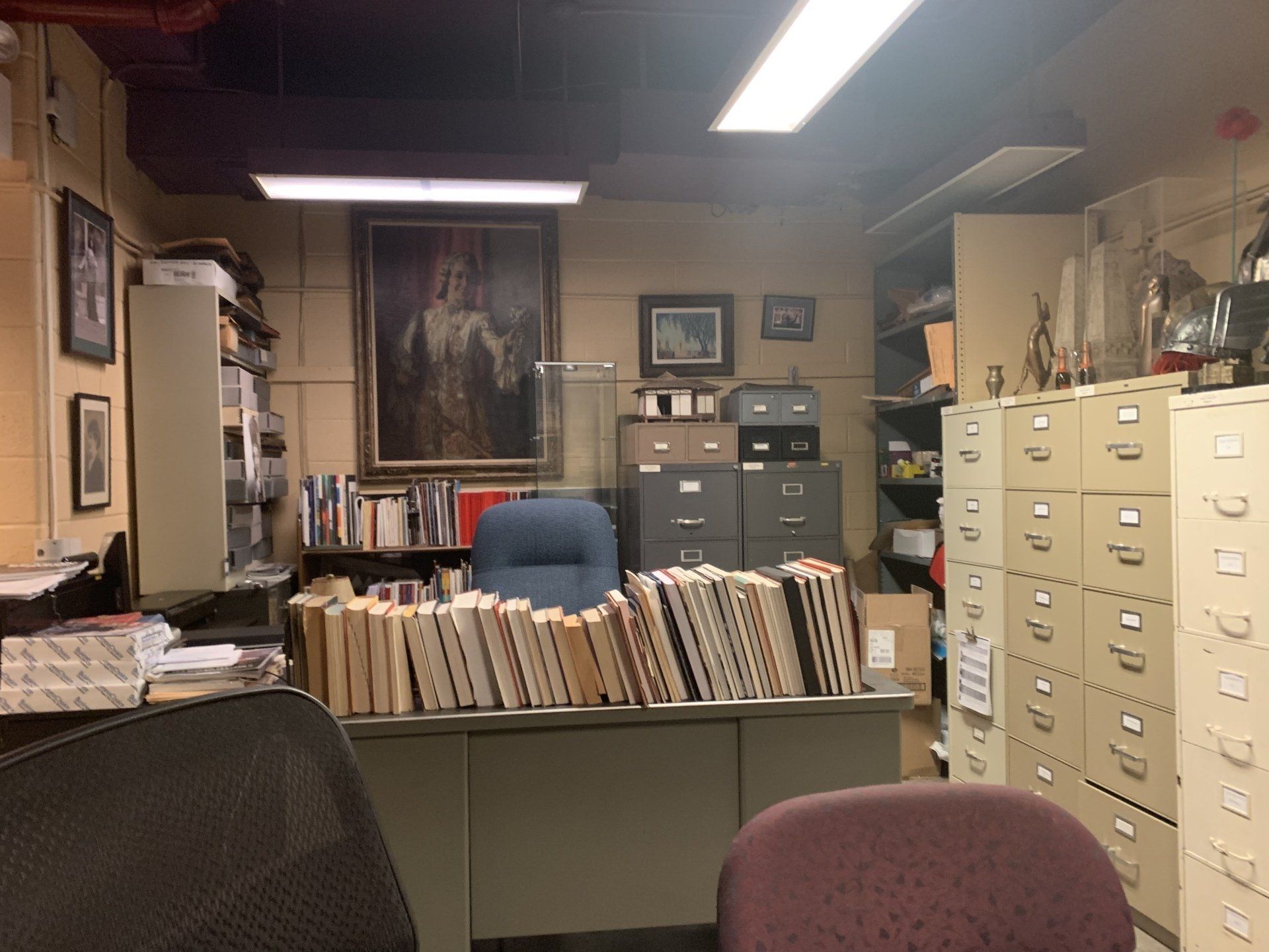 Cluttered office with desk, books, file cabinets, and a portrait on the wall.