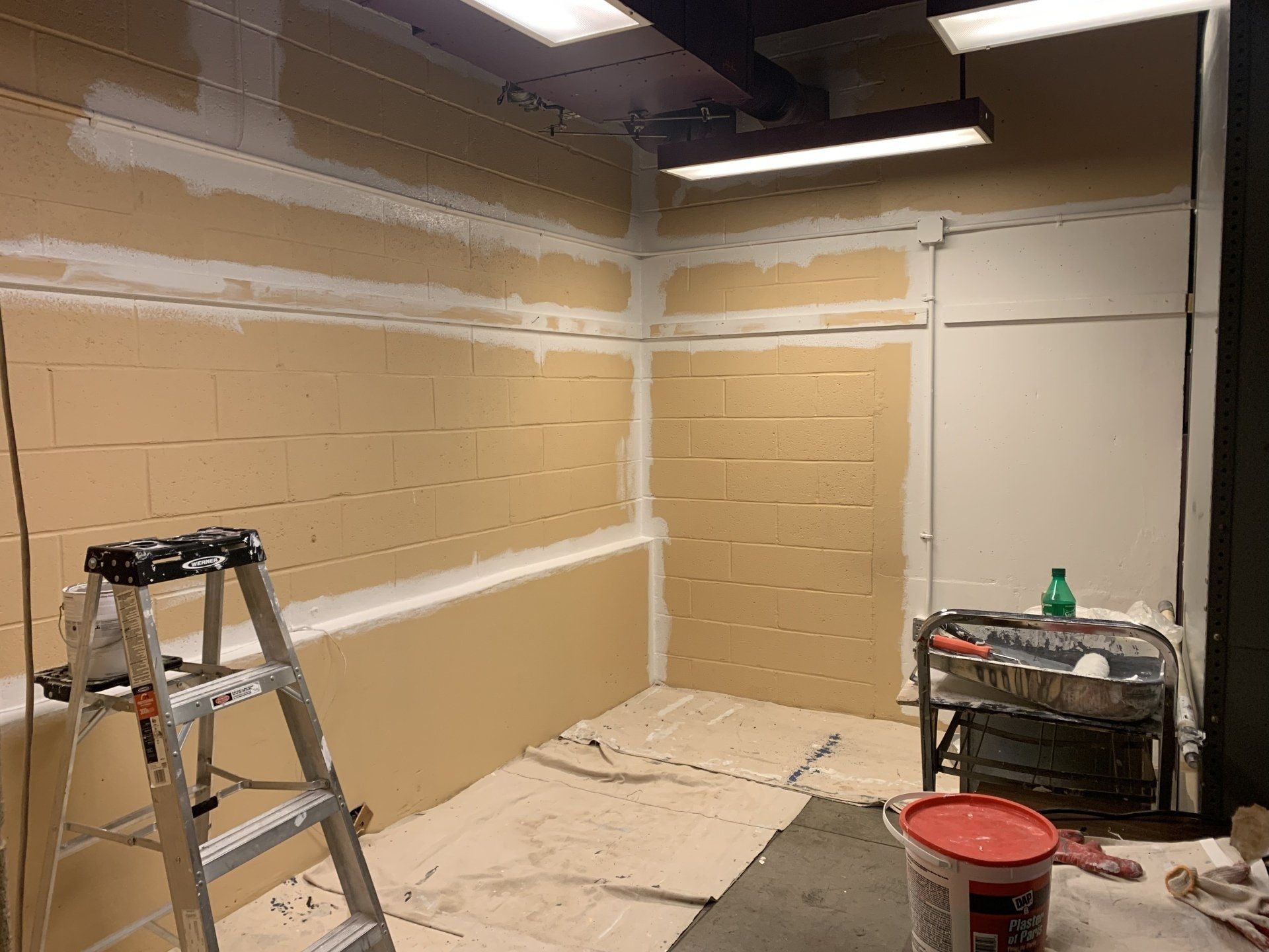 Room under renovation; tan brick walls, white drywall, ladder, paint cans, and tools present.