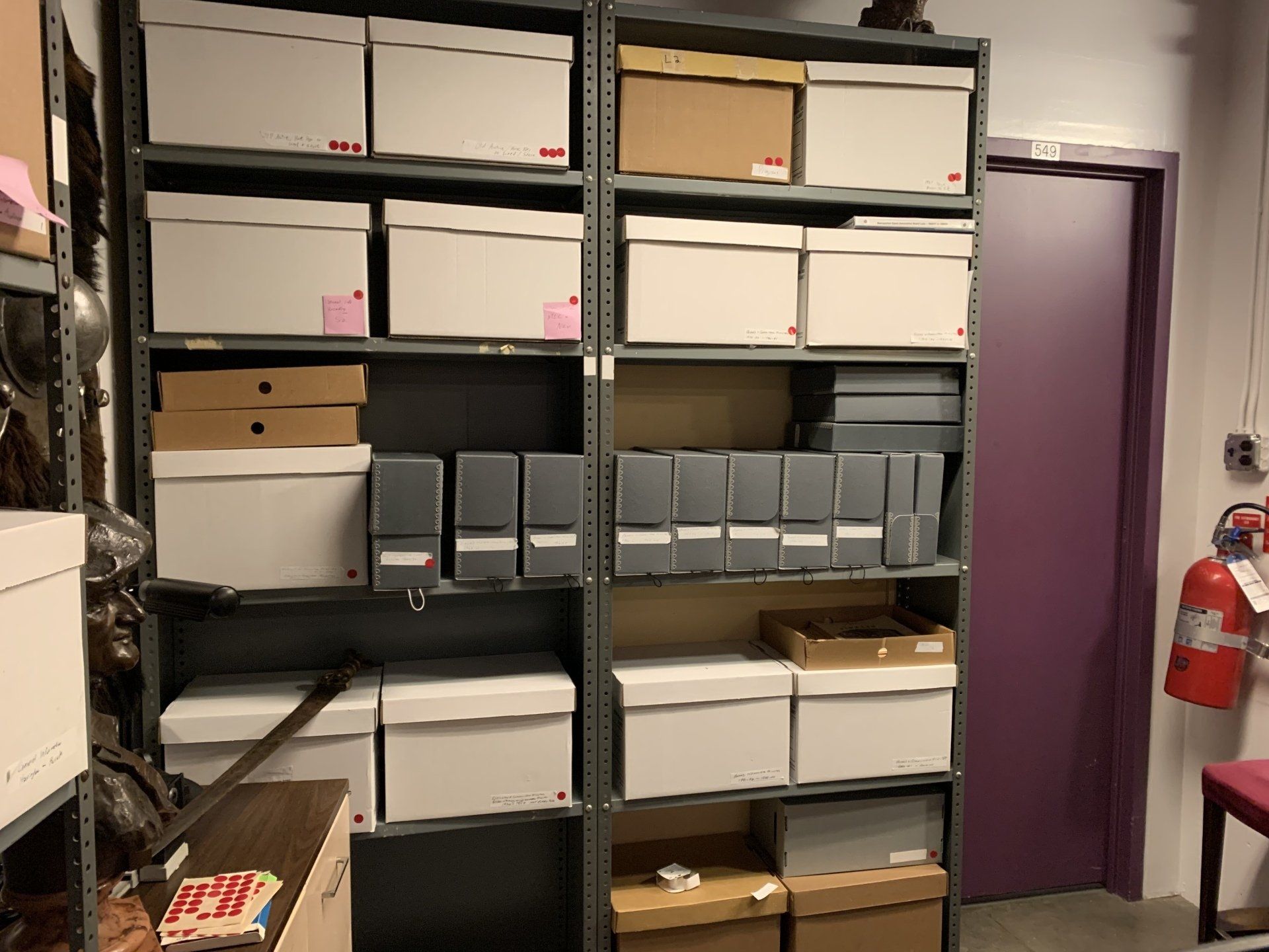 Shelves filled with white and tan boxes, file folders, and other materials in a storage room; a purple door is visible.