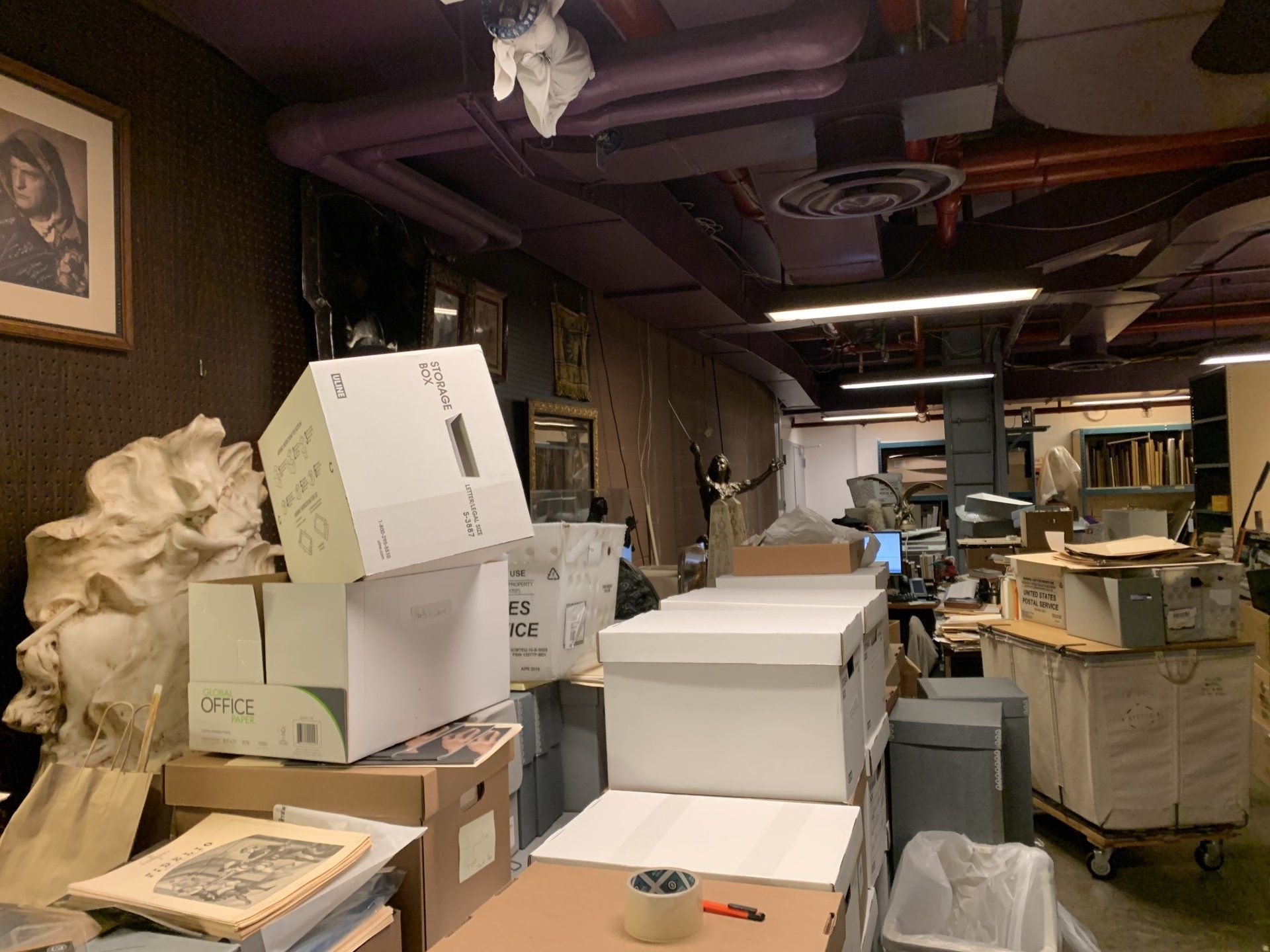 Boxes and clutter in a storage room with a painting on the wall and a cart.
