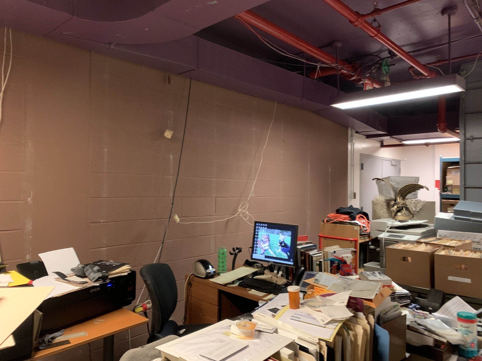 Cluttered office workspace with computer, desk, boxes, papers, and wires; pink ceiling, beige wall.