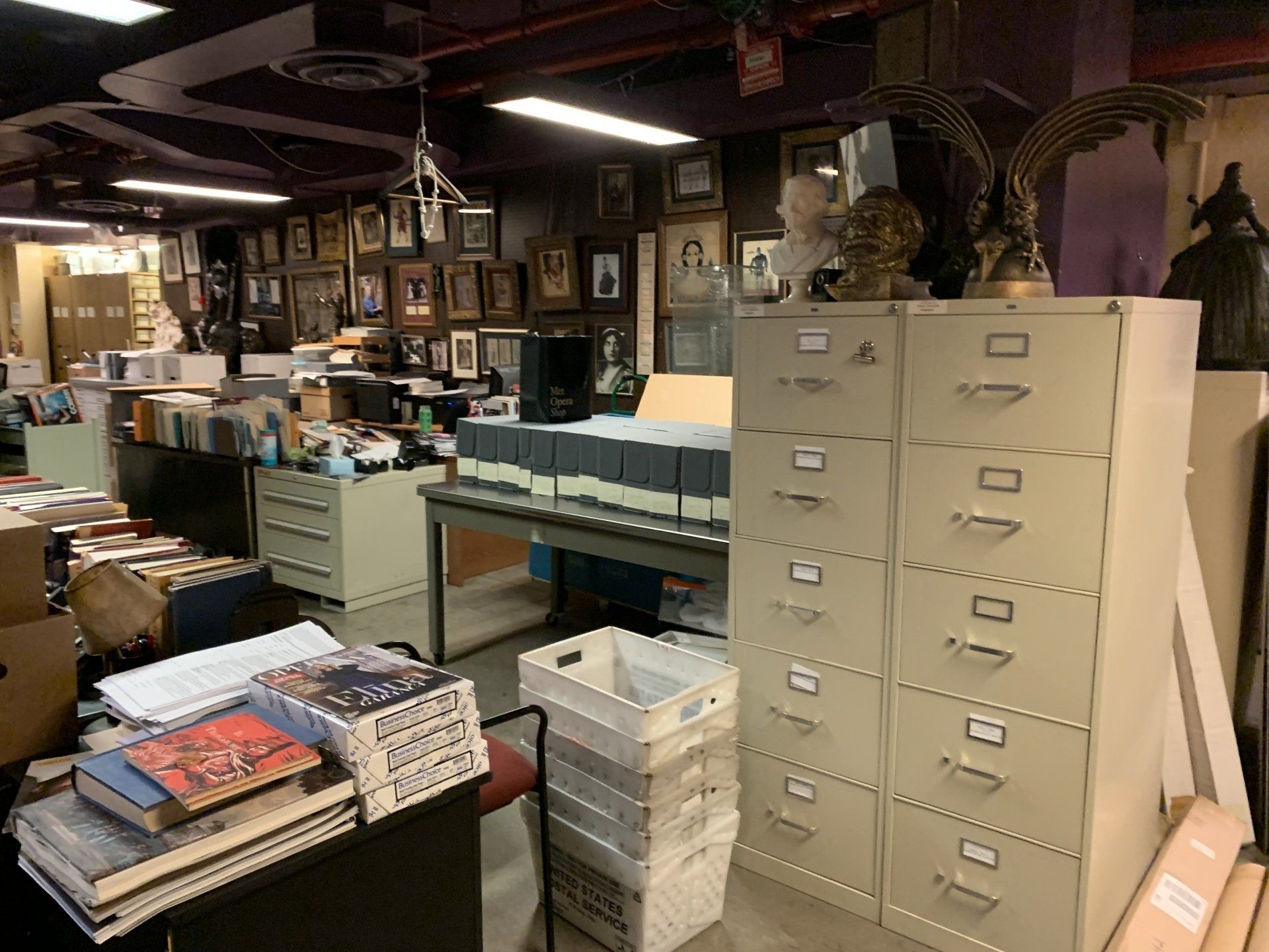 Cluttered office: filing cabinets, tables with stacks of books, framed photos on walls.