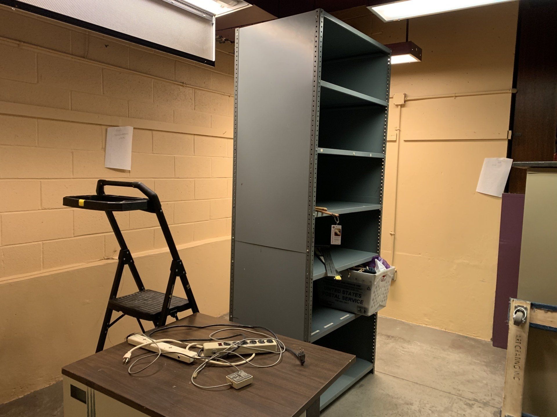 Tall gray shelving unit, desk with wires and a laptop, and a folding ladder in a room.
