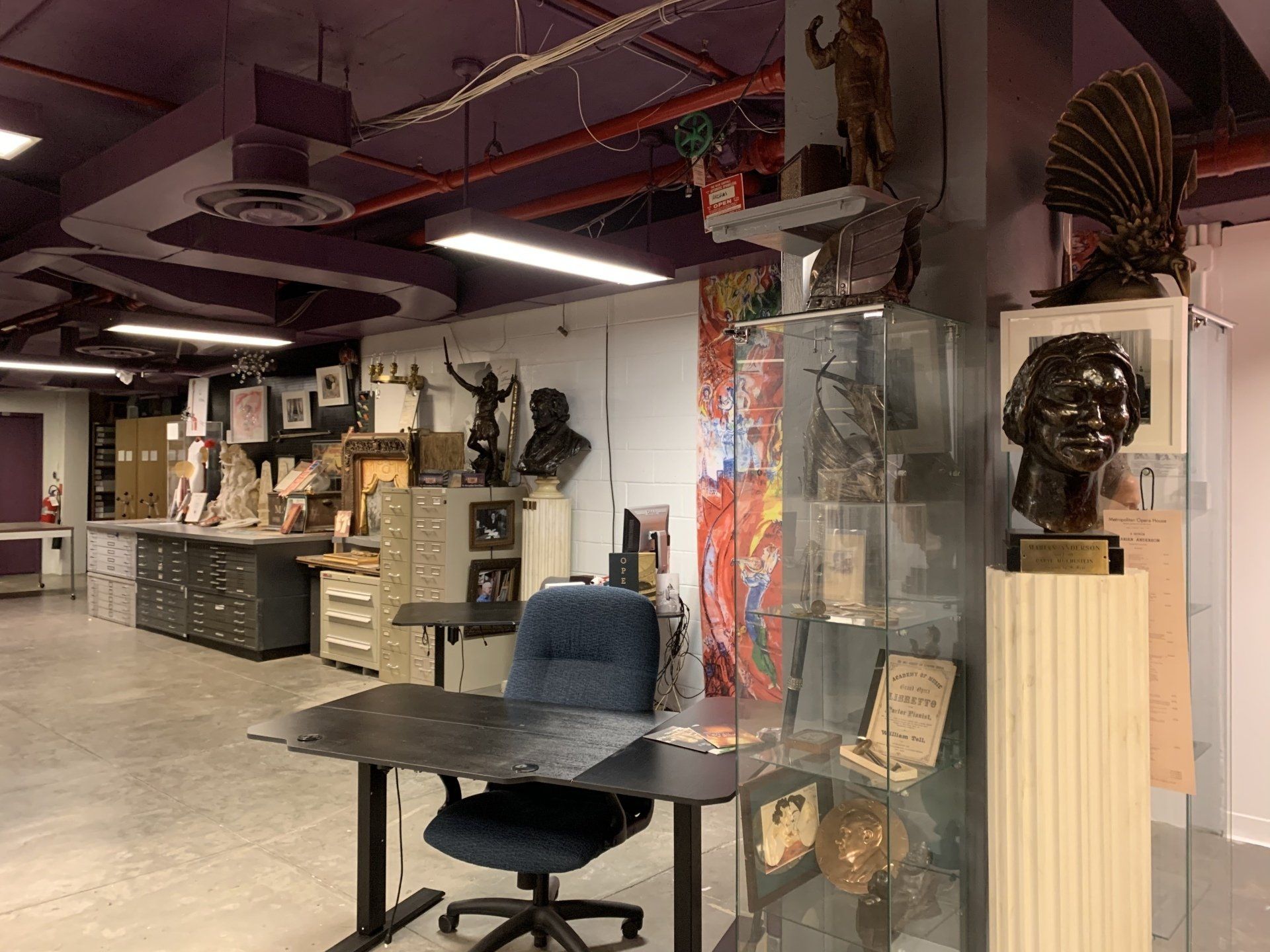 An art studio with sculptures, paintings, filing cabinets, and a desk. The room has purple accents.