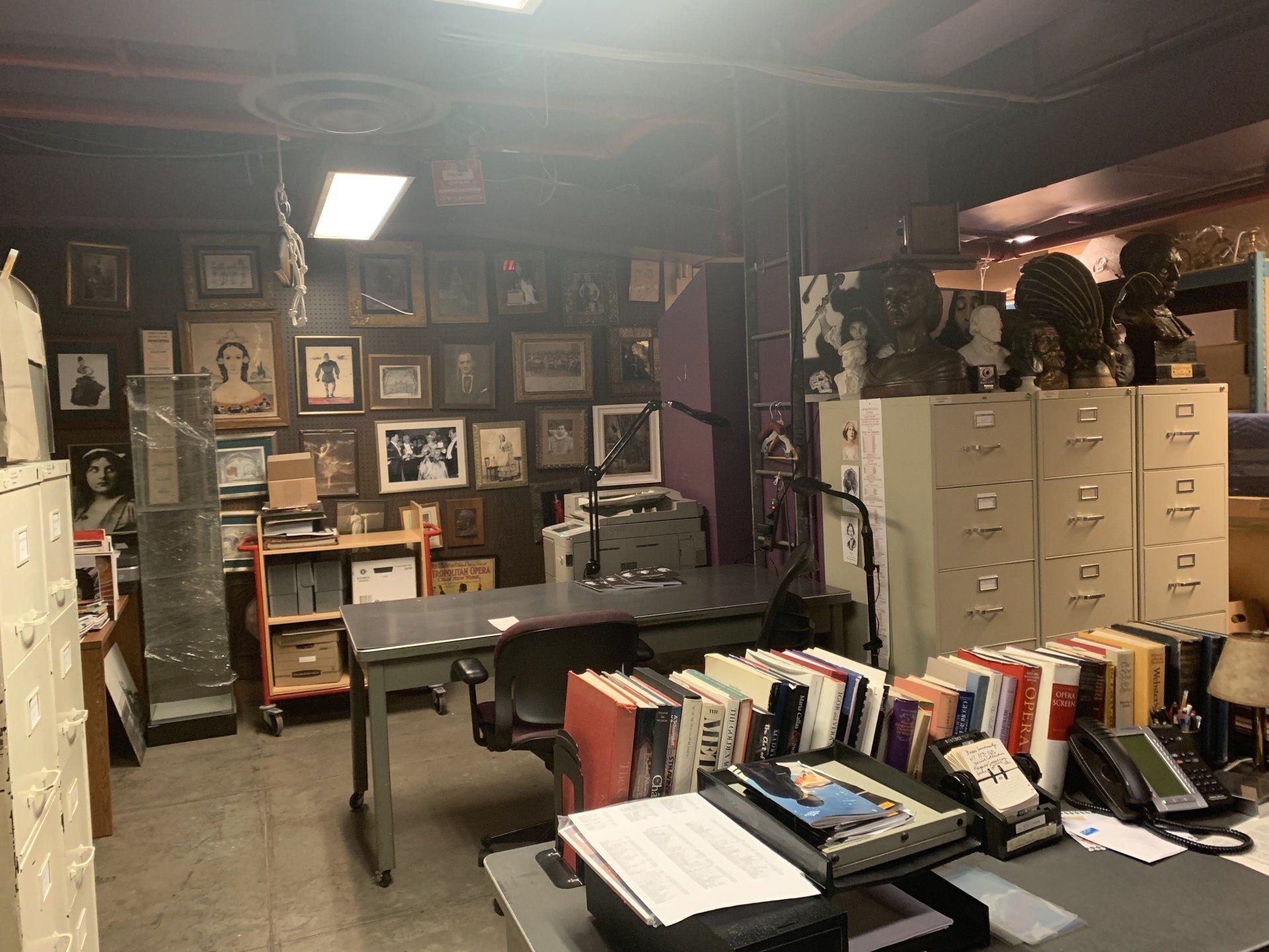 Messy office with many framed photos on the wall, filing cabinets, and a desk covered in papers and books.