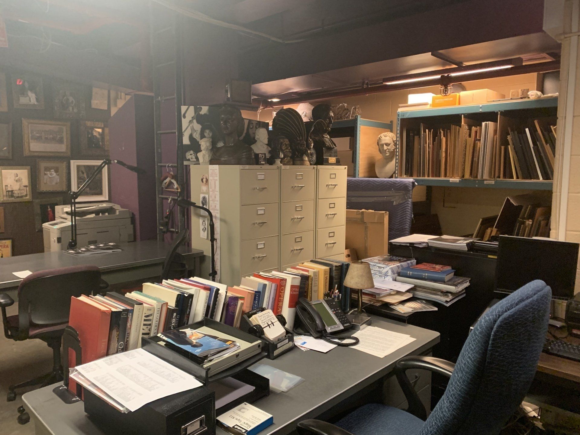 Cluttered office space with filing cabinets, desks, books, and artwork.