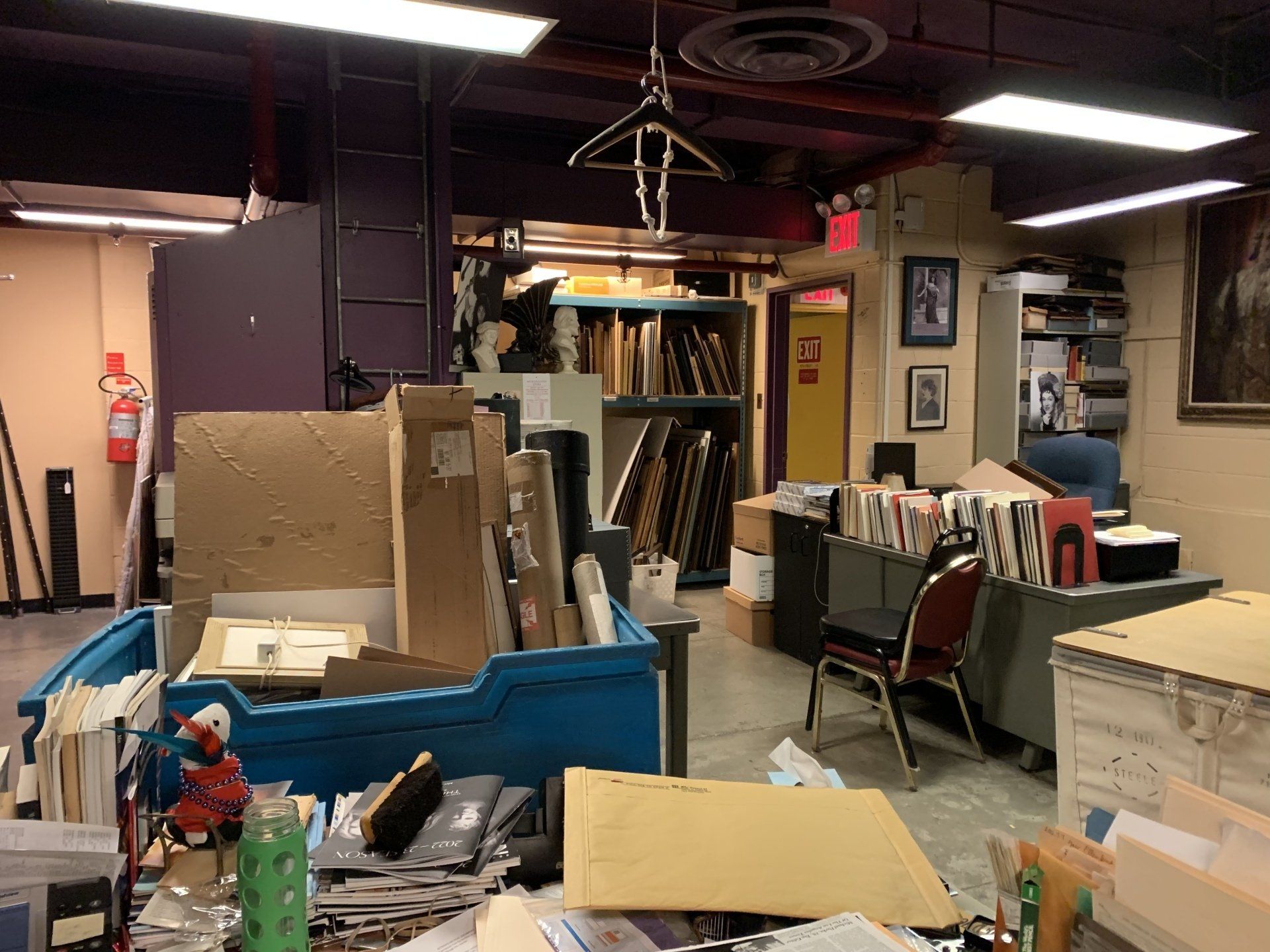 Cluttered art storage room: boxes, frames, files, sculptures, a blue bin, and a fire extinguisher.
