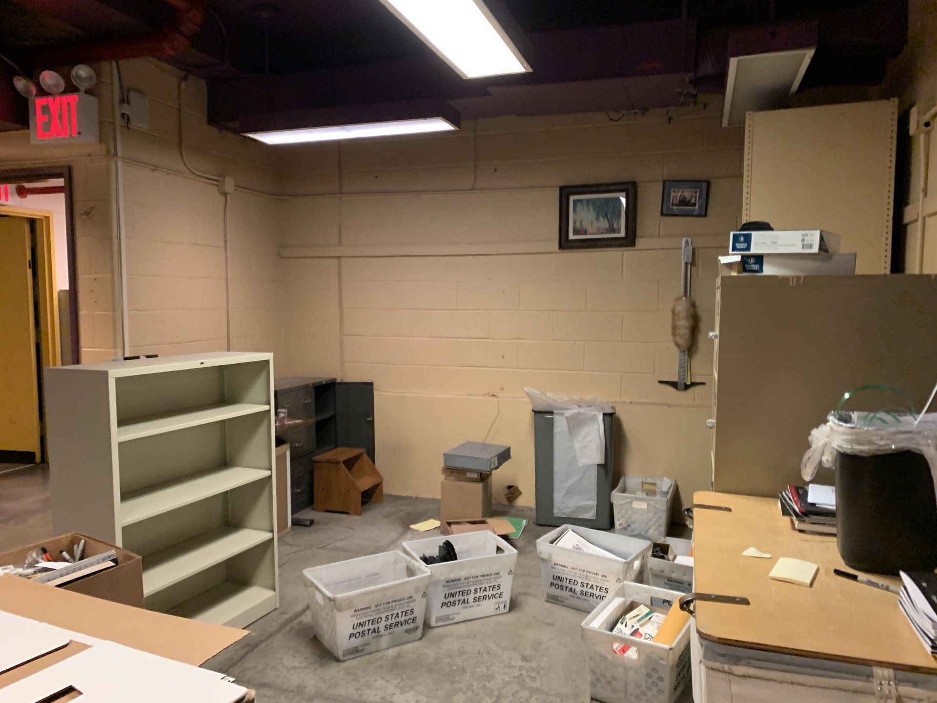 Basement office: shelves, boxes, desk with items, old vacuum, beige walls, fluorescent lighting.