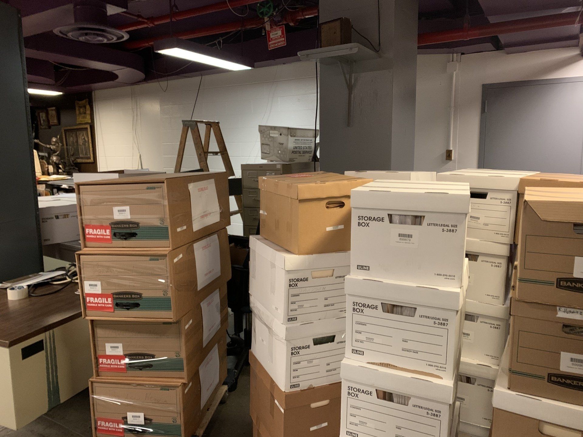 Boxes stacked in an office storage room. Beige, white boxes fill the frame. A small wooden step ladder is also present.