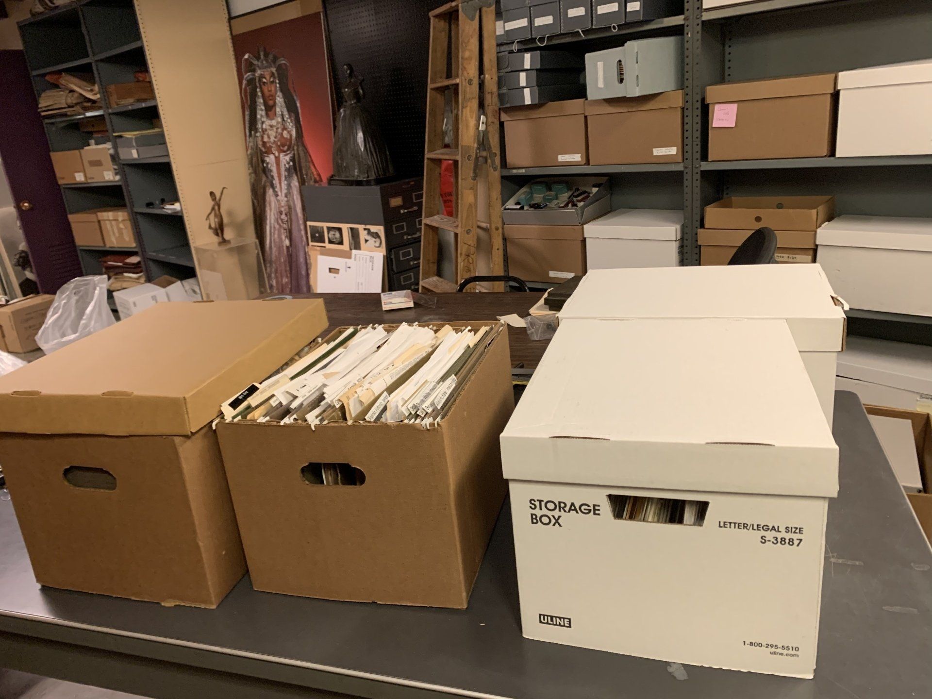 Three cardboard boxes on a table; two tan, one white. Boxes are open, some papers visible. Shelves with boxes are in the background.