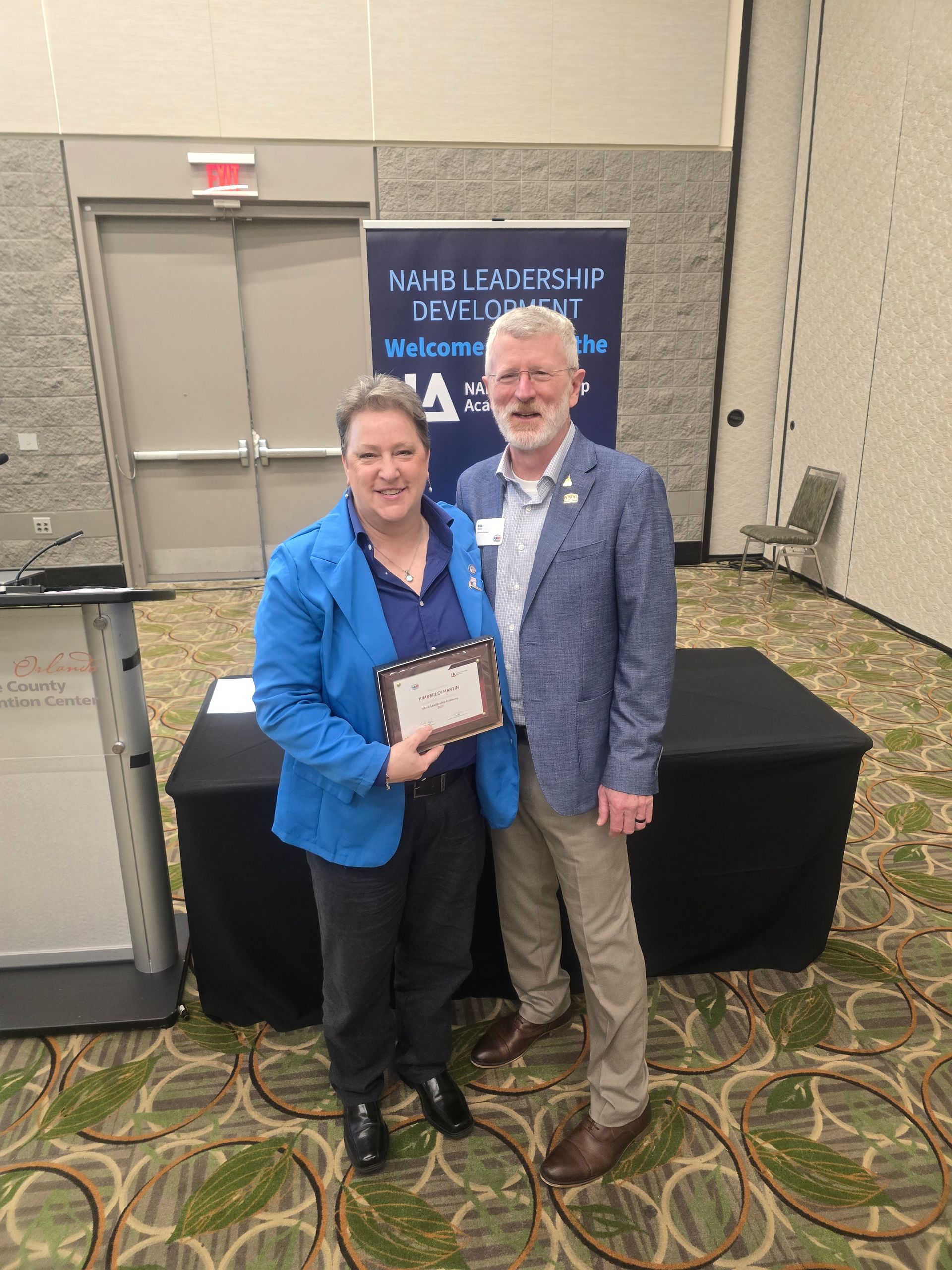 Two people pose for a photo with a plaque at a conference; a woman in a blue jacket and a man in a blazer smile.