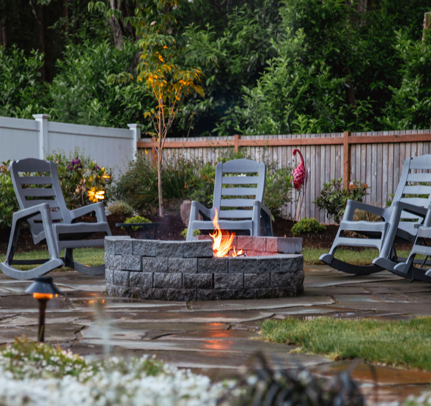 A fire pit in a backyard with rocking chairs around it