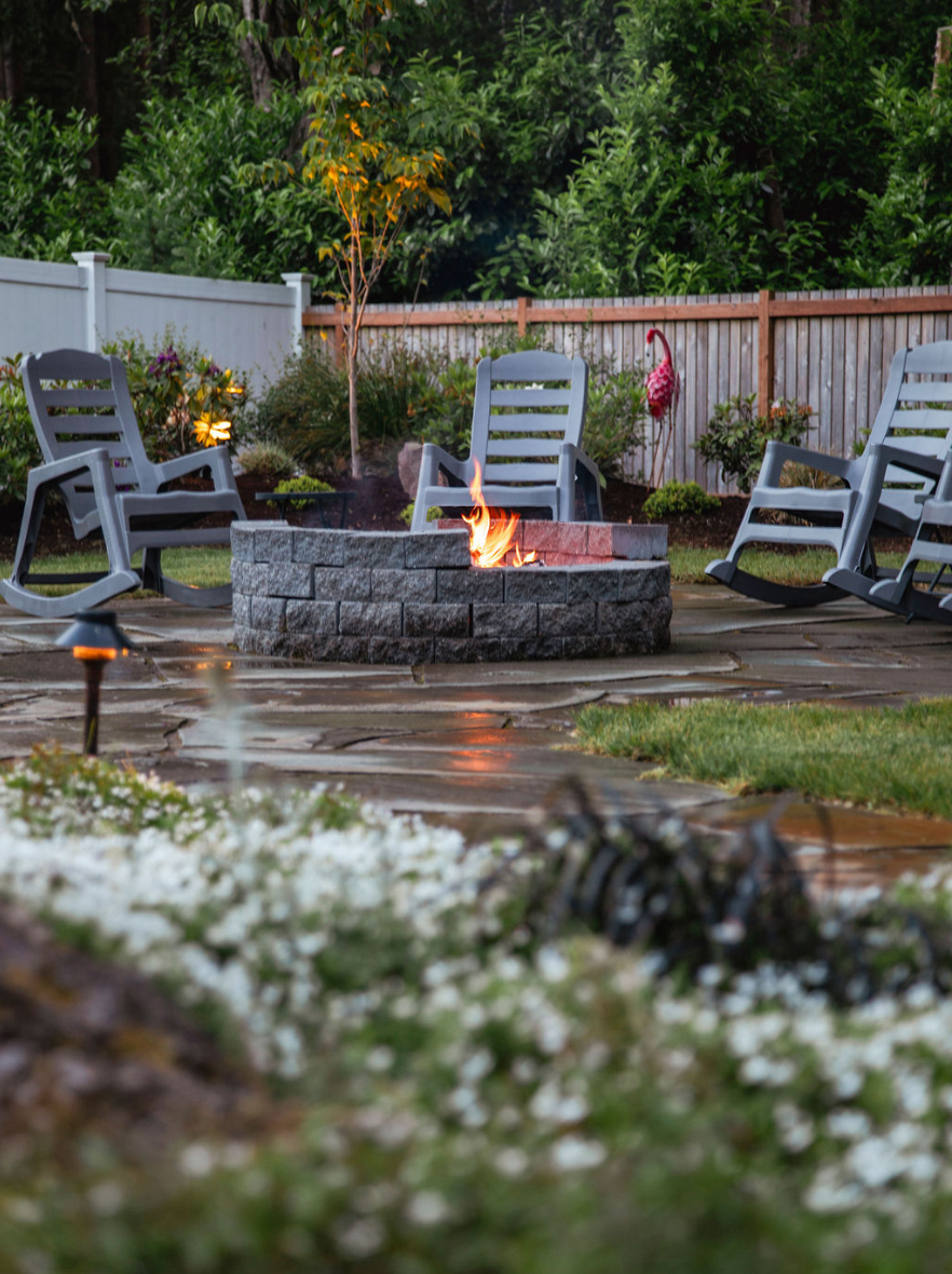 There is a fire pit in the middle of the backyard surrounded by rocking chairs.