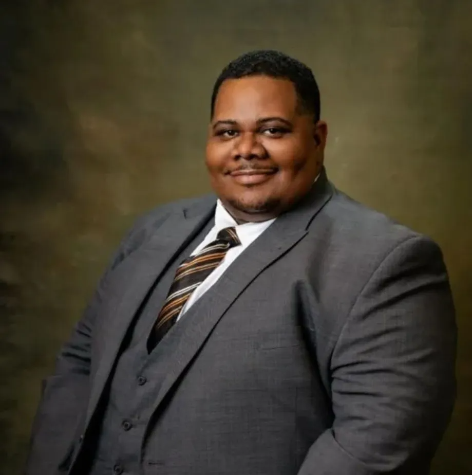 Studio portrait of a smiling man in a gray suit and striped tie against a blurred brown background