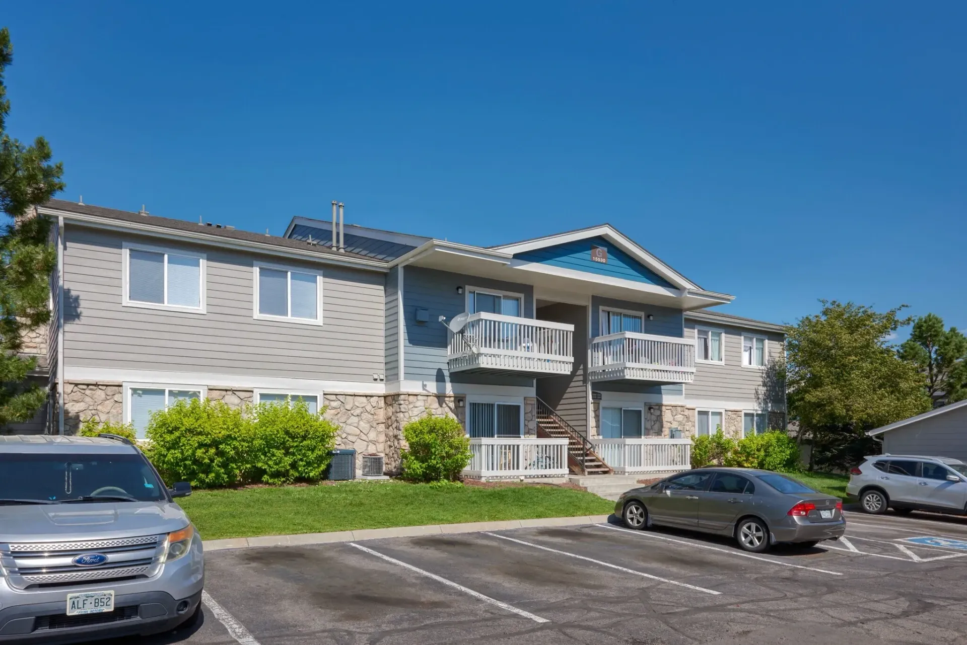 Apartment building with balconies and parked cars in a sunny lot under a blue sky
