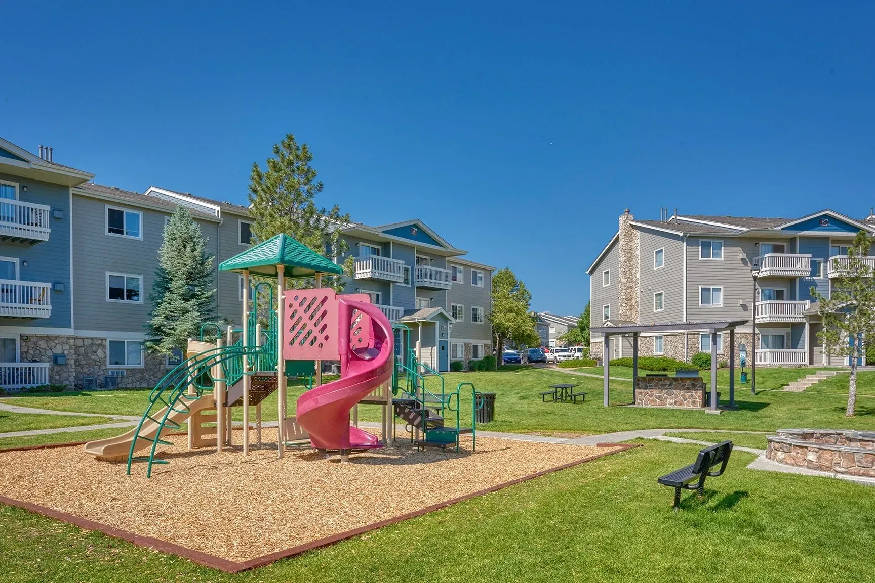 Apartment complex playground with pink slide, green climbing frame, benches, and grassy courtyard under a blue sky