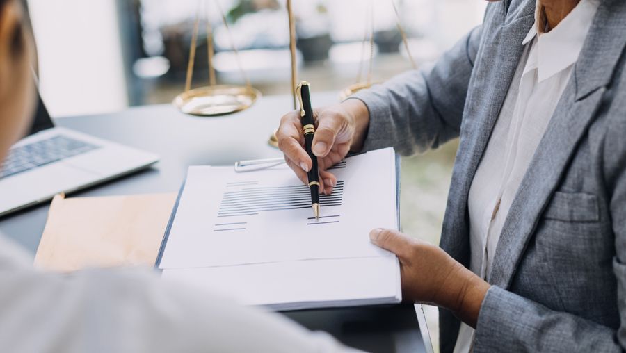 A professional sitting at a desk holds a pen and points to a document while consulting with another person.
