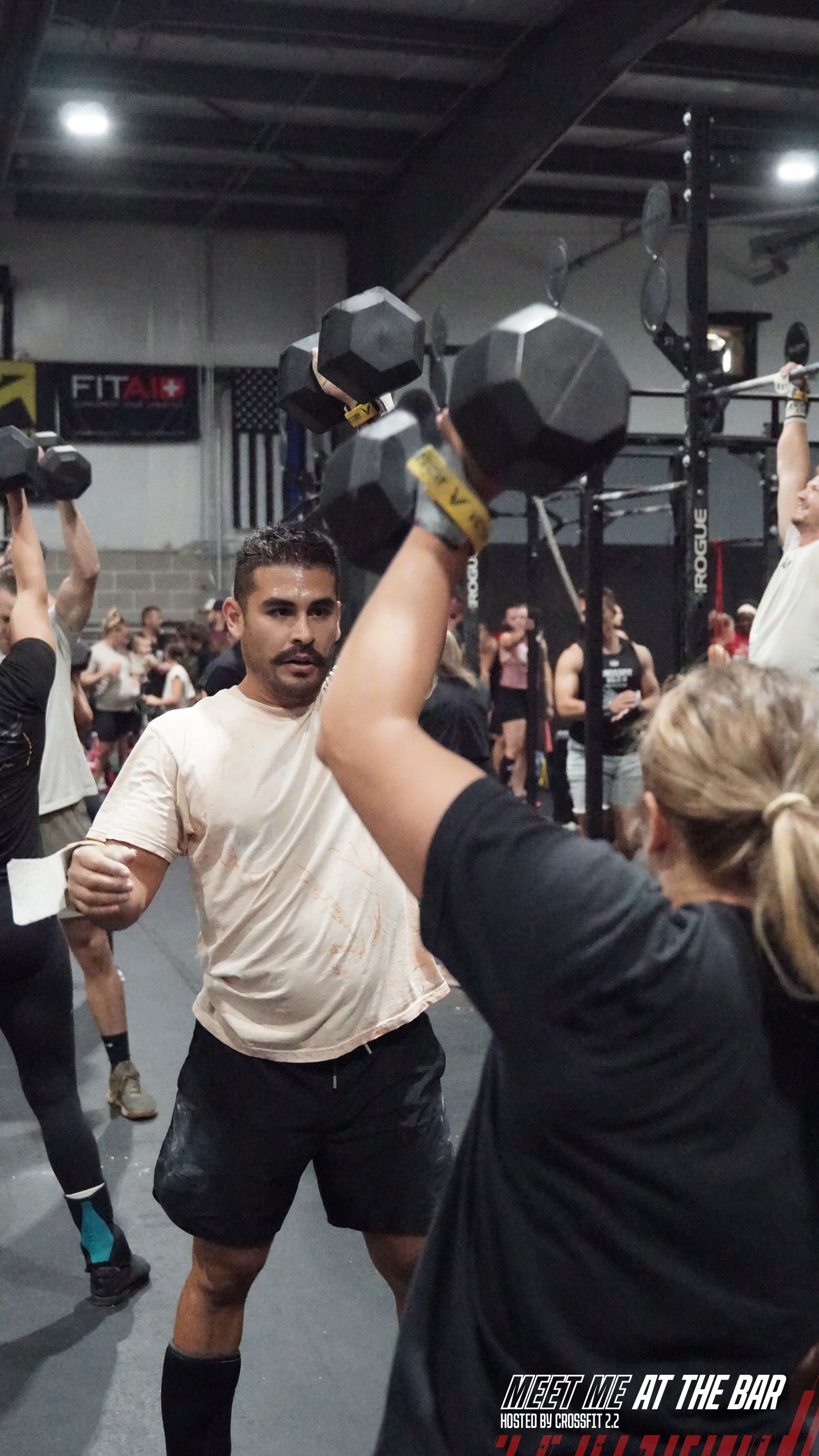Man jumps onto a wooden box at a gym with others working out, and a barbell is held overhead.